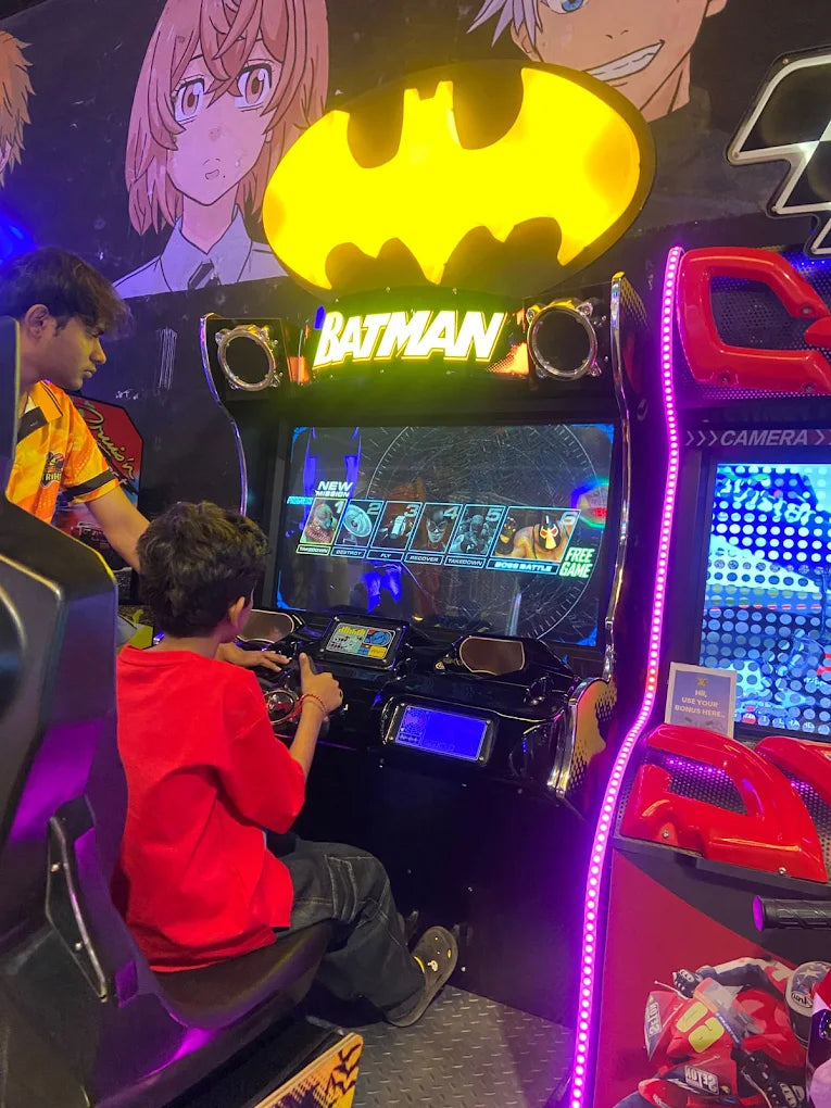 Children play a Batman arcade game in a dimly lit arcade with anime-style murals.