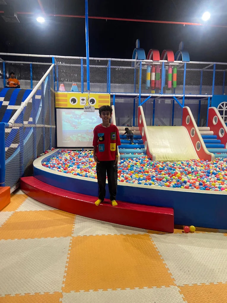 A young boy stands in a ball pit at an indoor playground. He is smiling in front of a colorful slide and a screen.