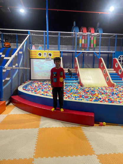 A young boy stands in a ball pit at an indoor playground. He is smiling in front of a colorful slide and a screen.