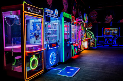 A row of brightly lit arcade games, including claw machines and video games, in a dark arcade.