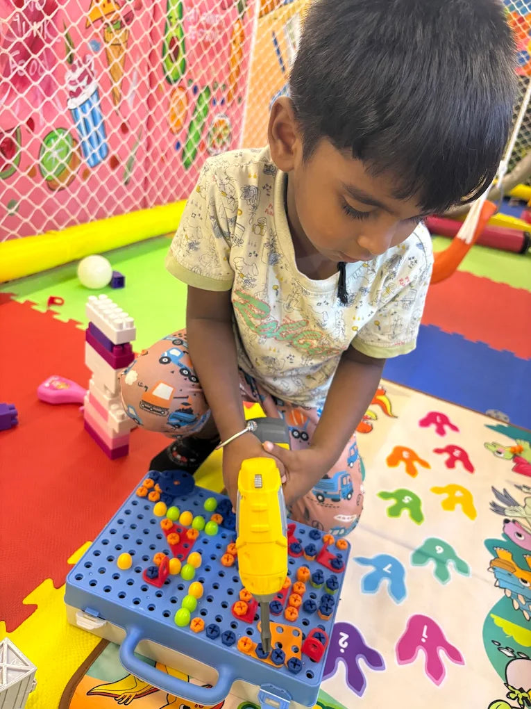 A young child plays with a toy drill and building set on a colorful mat in a playroom.