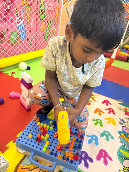 A young child plays with a toy drill and building set on a colorful mat in a playroom.