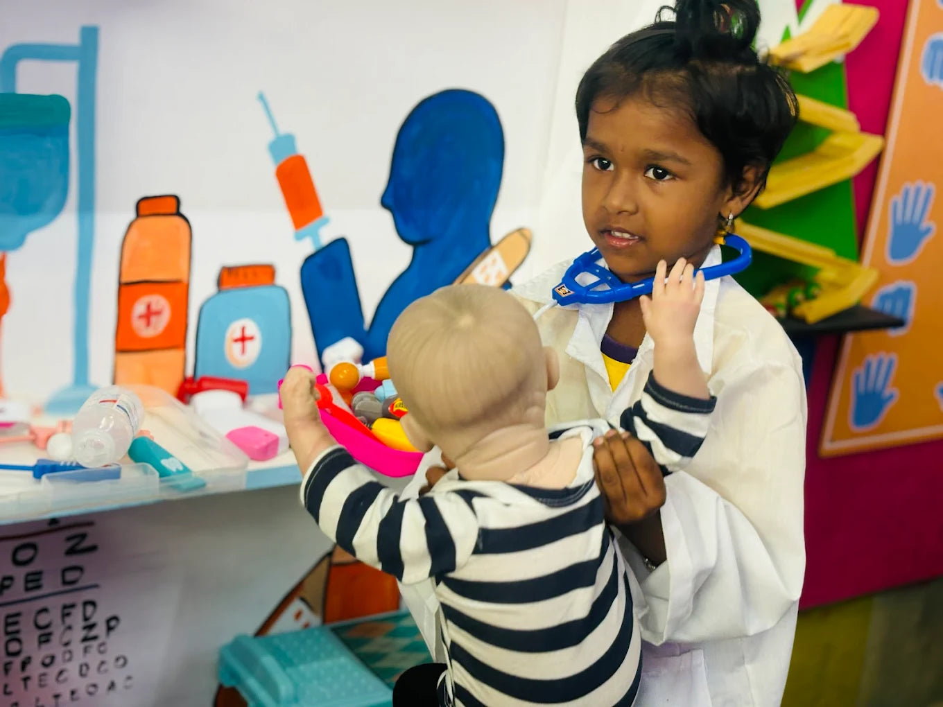A young girl in a doctor's coat plays doctor with a baby doll in a room with medical-themed decorations.