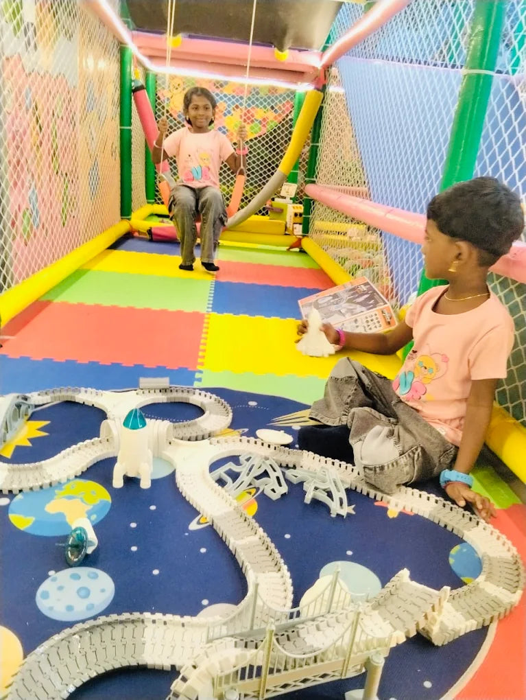 Two young children play in a colorful indoor playground with a toy train set.