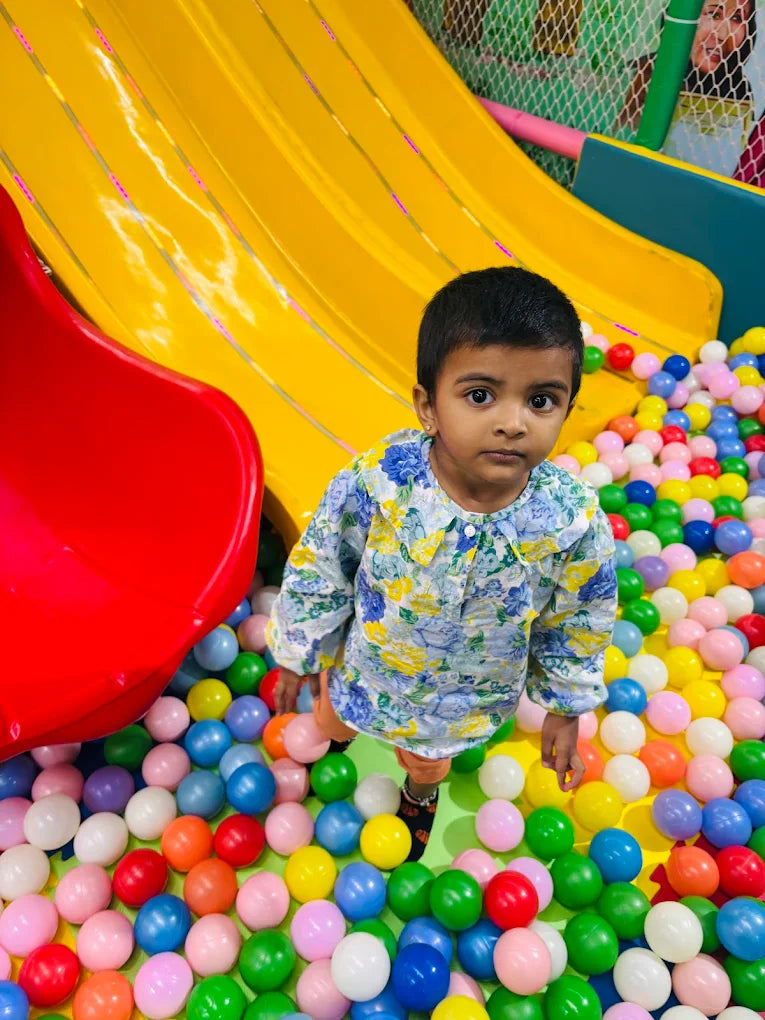 A young child stands in a ball pit next to a yellow slide.