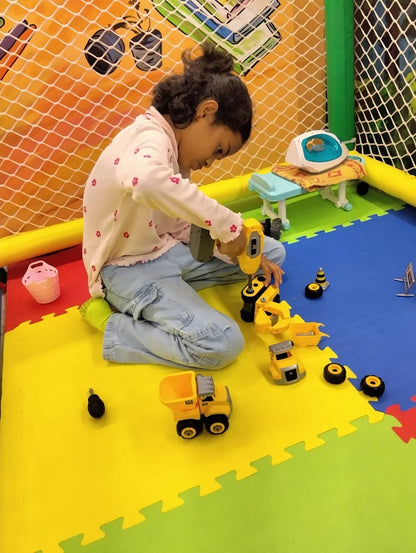 A young child plays with toy construction vehicles on a colorful mat in a play area.