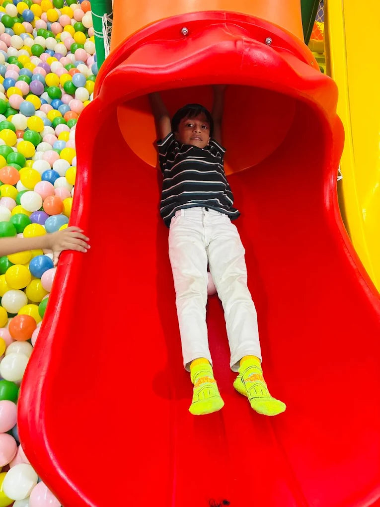 A young boy slides down a red tube slide into a ball pit. He is wearing white pants and bright yellow socks.