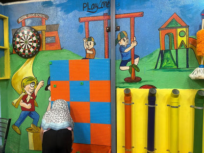Children's play area with a mural of a school and playground, featuring a dartboard and colorful tubes.