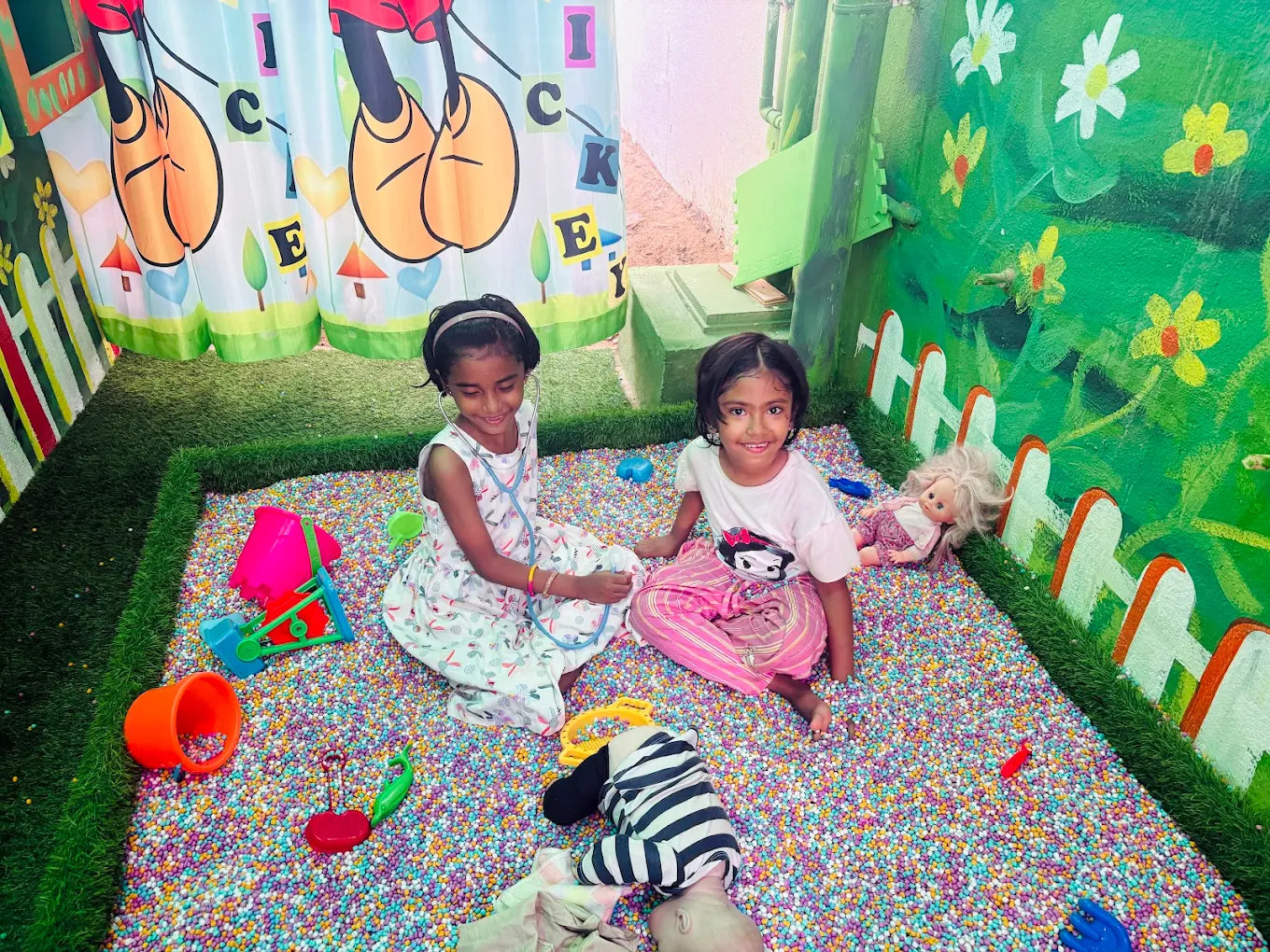 Two young girls play in a ball pit with toys, one wearing a stethoscope.