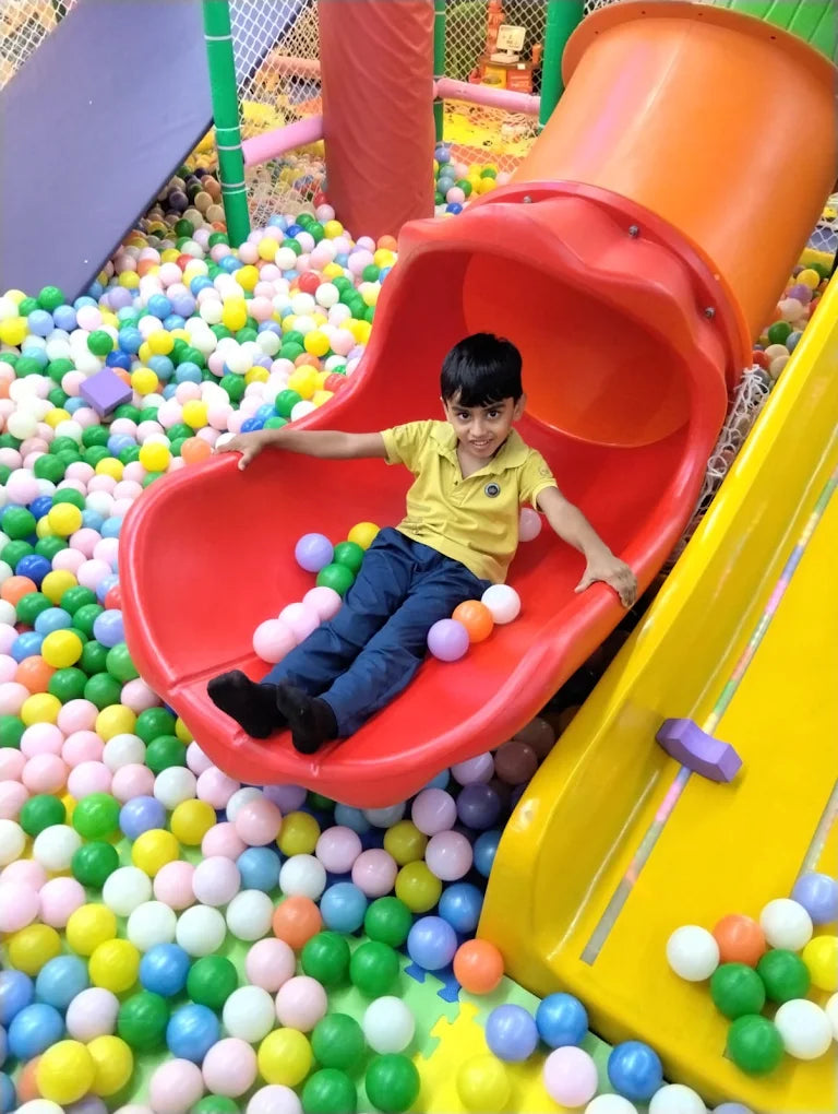 A young boy smiles as he sits on a red slide in a ball pit filled with colorful balls.