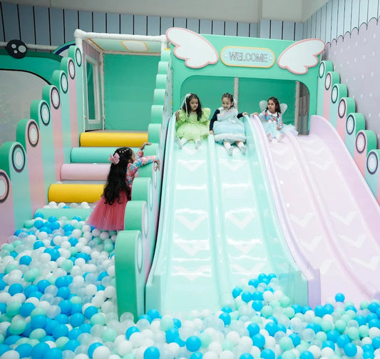 Young girls in dresses play on a colorful indoor playground with slides and a ball pit.