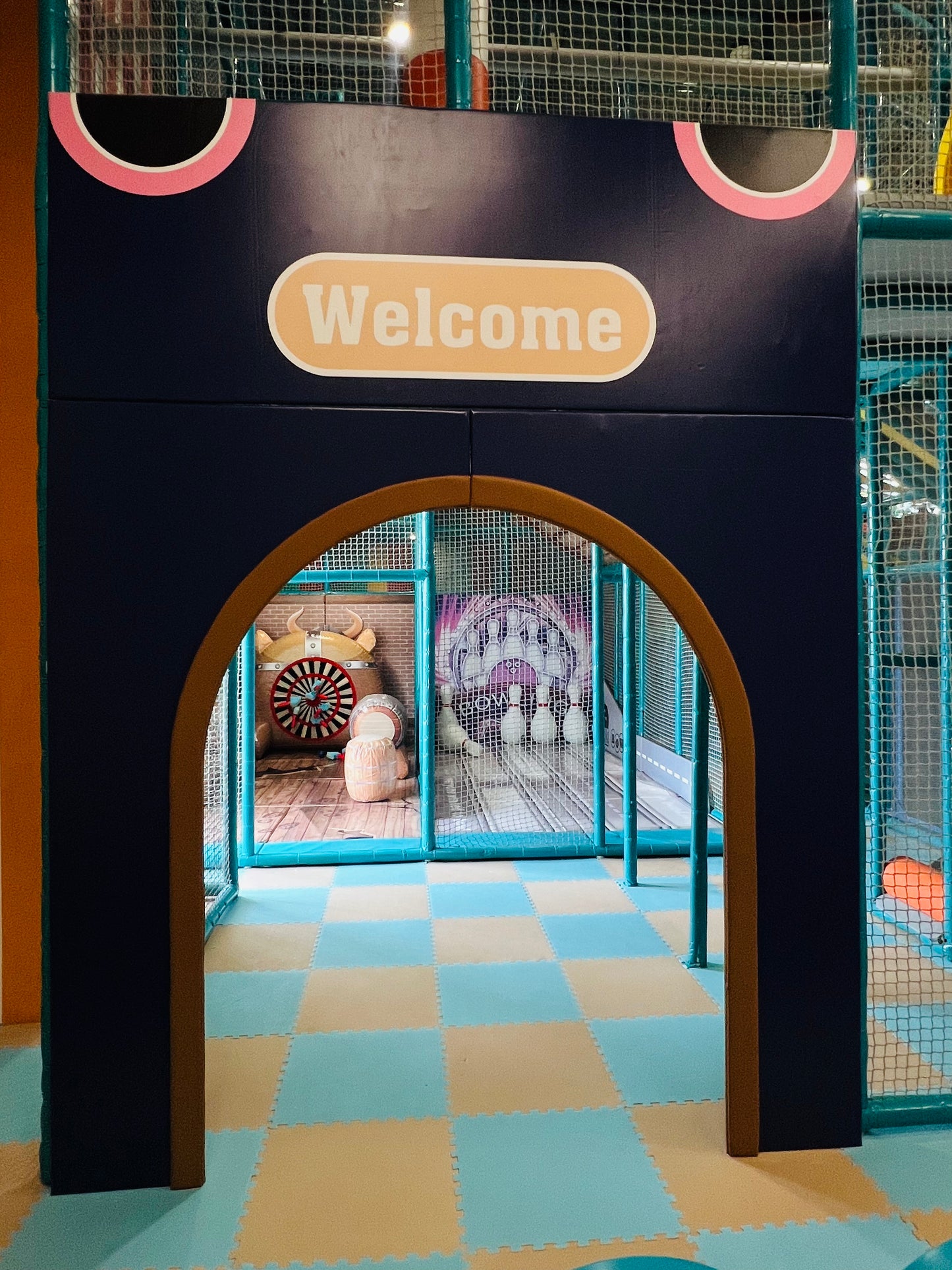 An arched entryway to a children's play area with a "Welcome" sign. Inside, a bowling game and other play structures are visible.