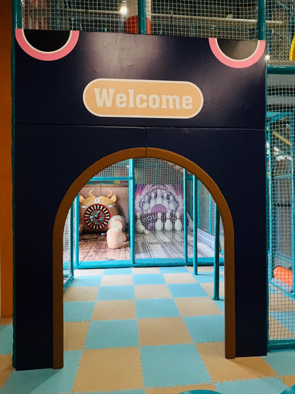 An arched entryway to a children's play area with a "Welcome" sign. Inside, a bowling game and other play structures are visible.