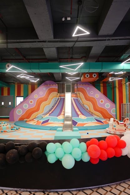 A colorful indoor playground with a large slide and soft play area, decorated with balloons.
