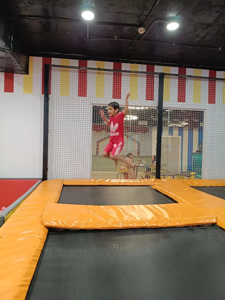 A young boy in a red shirt and shorts jumps high on a trampoline. He is in an indoor play area with netting and colorful decorations.