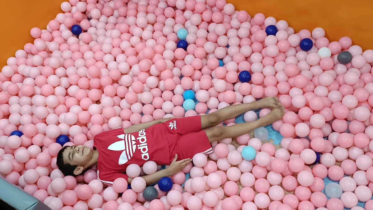 A young boy lies in a ball pit filled with pink and blue balls, wearing a red Adidas outfit.