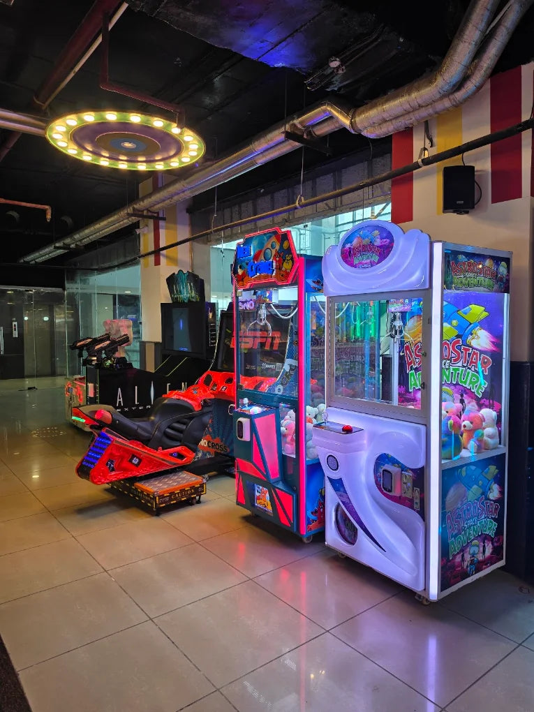 Arcade with various games, including a motorcycle simulator and claw machines, lit by neon lights.