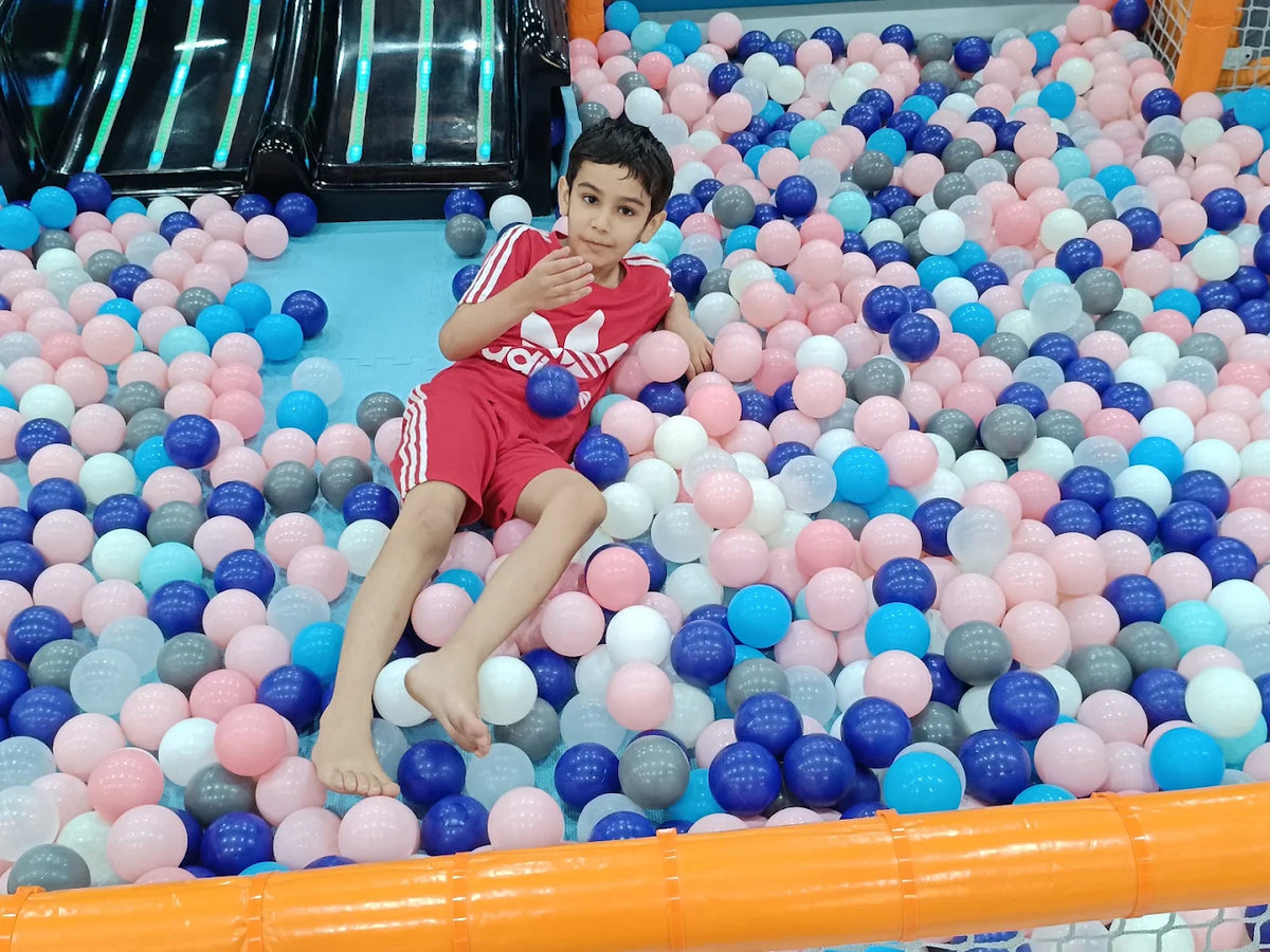 A young boy in a red shirt and shorts sits in a ball pit filled with pink, blue, and white balls.