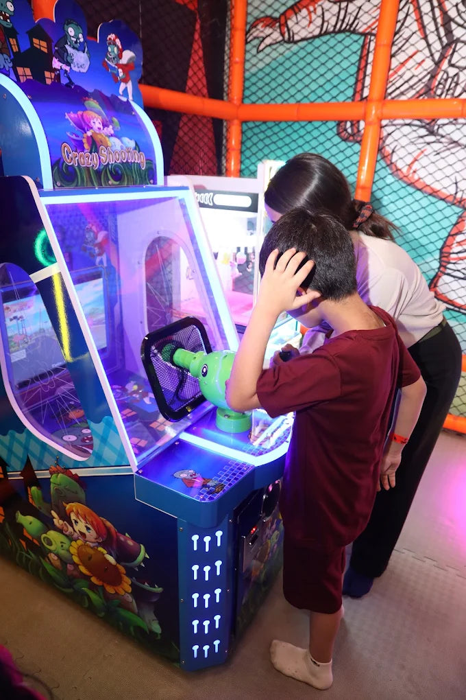 A child plays a "Crazy Shooting" arcade game with a woman standing behind them. The game features cartoon zombies and plants.