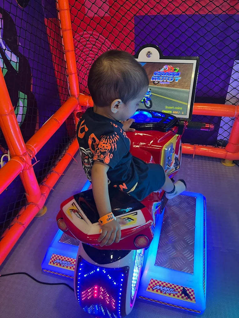 A young child plays a motorcycle arcade game in a colorful play area. The child is seated on the game, looking at the screen.