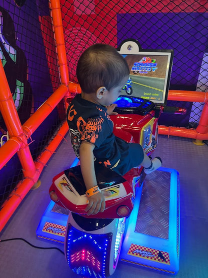 A young child plays a motorcycle arcade game in a colorful play area. The child is seated on the game, looking at the screen.