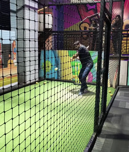 A man plays cricket indoors in a netted enclosure with a green floor and colorful walls.