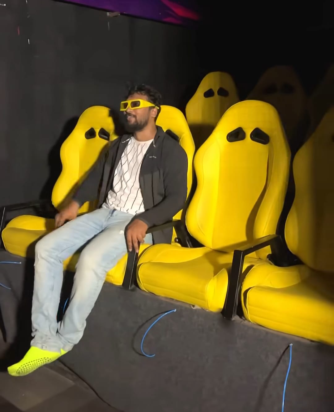 A young man wearing 3D glasses sits in a bright yellow seat in a dark room.