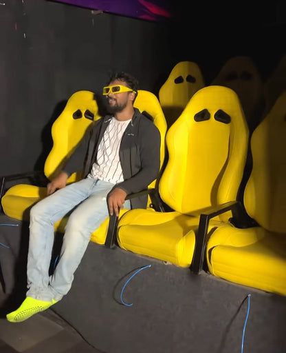 A young man wearing 3D glasses sits in a bright yellow seat in a dark room.