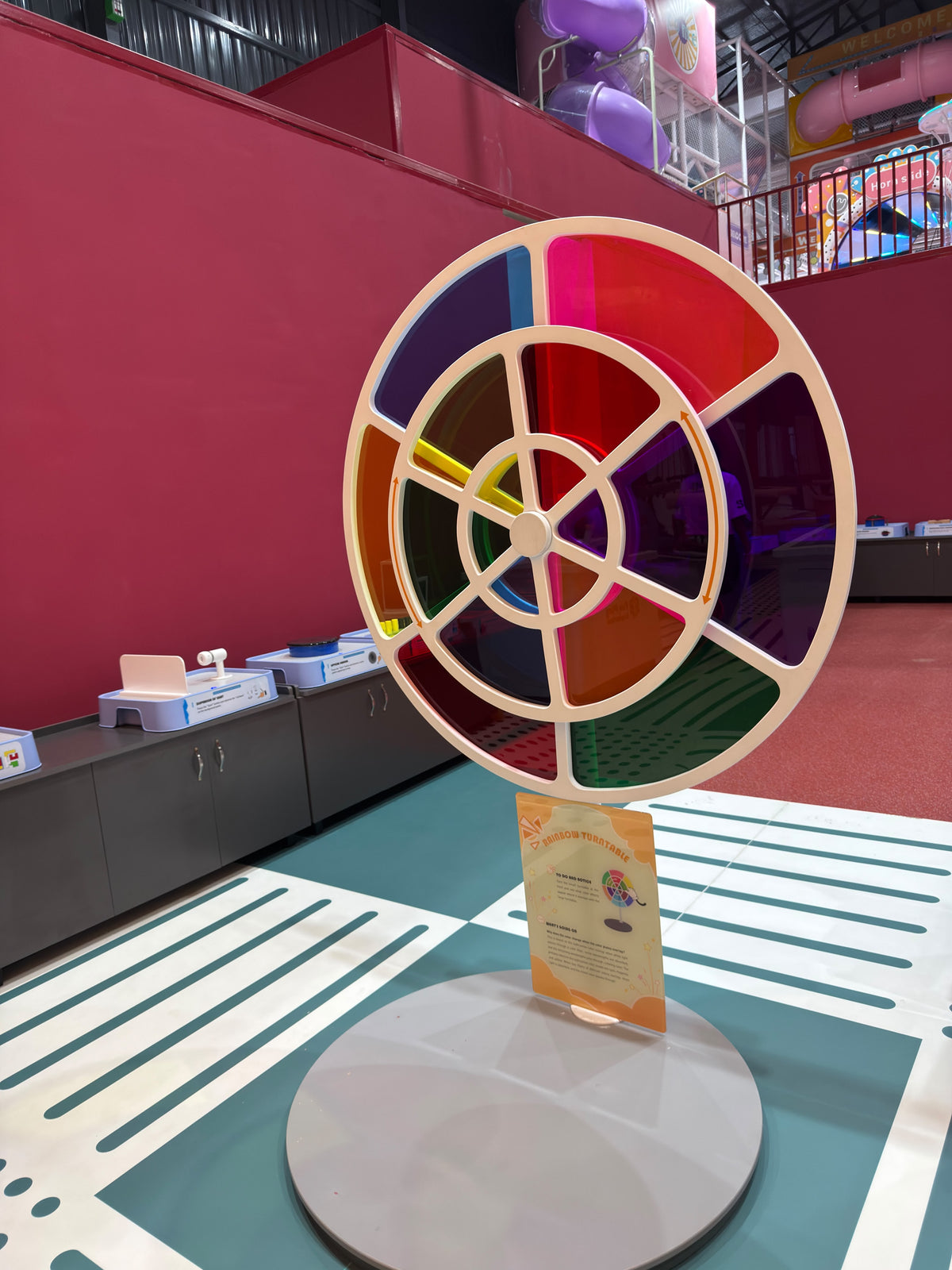 A colorful rainbow turntable is displayed in a children's play area with slides in the background.