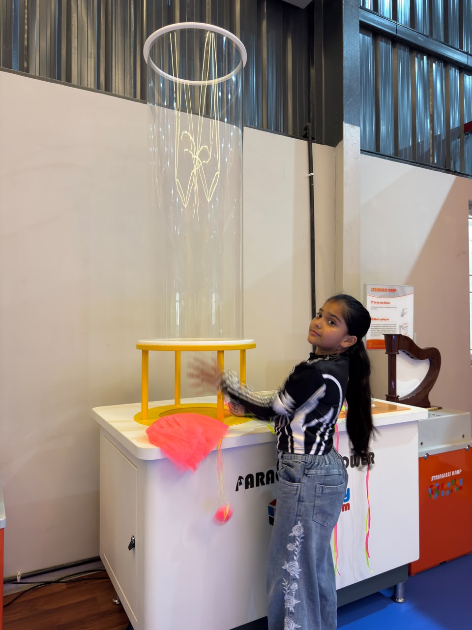 A young girl plays with a science exhibit featuring a large clear tube and a pink object. She is in a museum or science center.