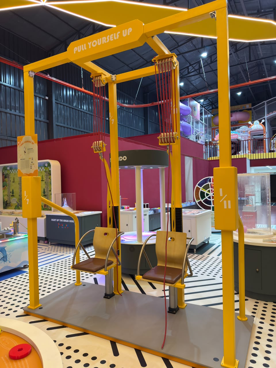 Children sit on a yellow pulley system at an indoor playground, ready to "pull yourself up."