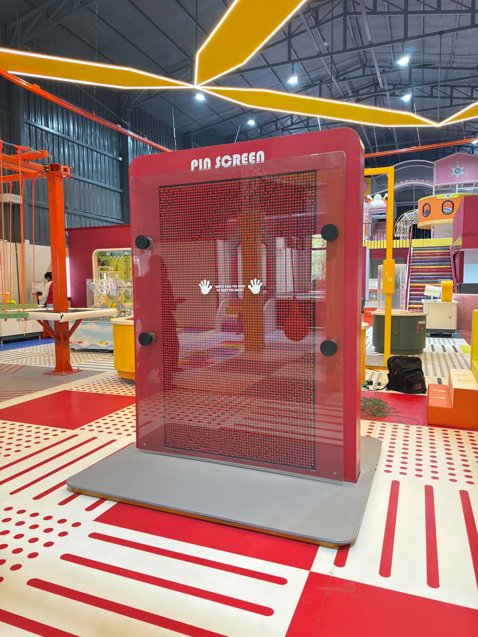 A large pin screen in a children's play area with colorful flooring and overhead lights.