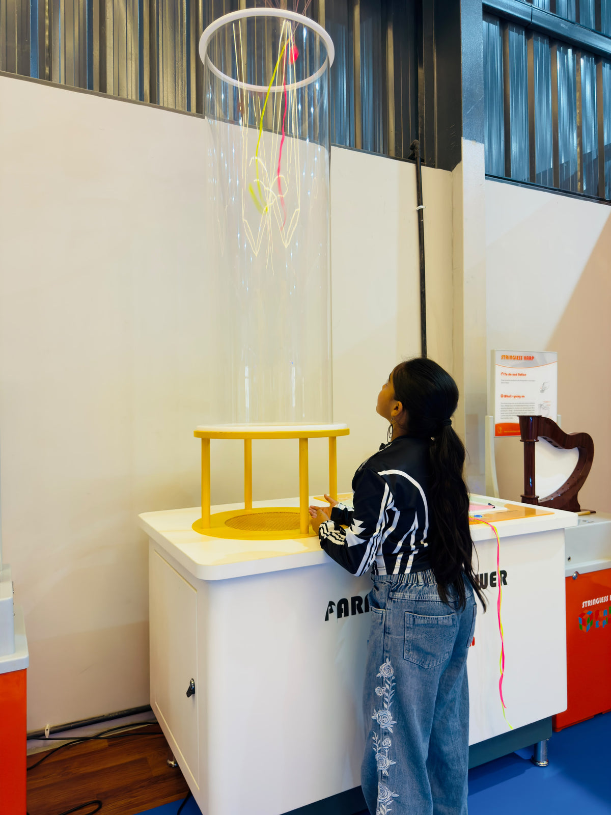 A young girl looks up at a clear tube with colorful streamers inside, at an exhibit.
