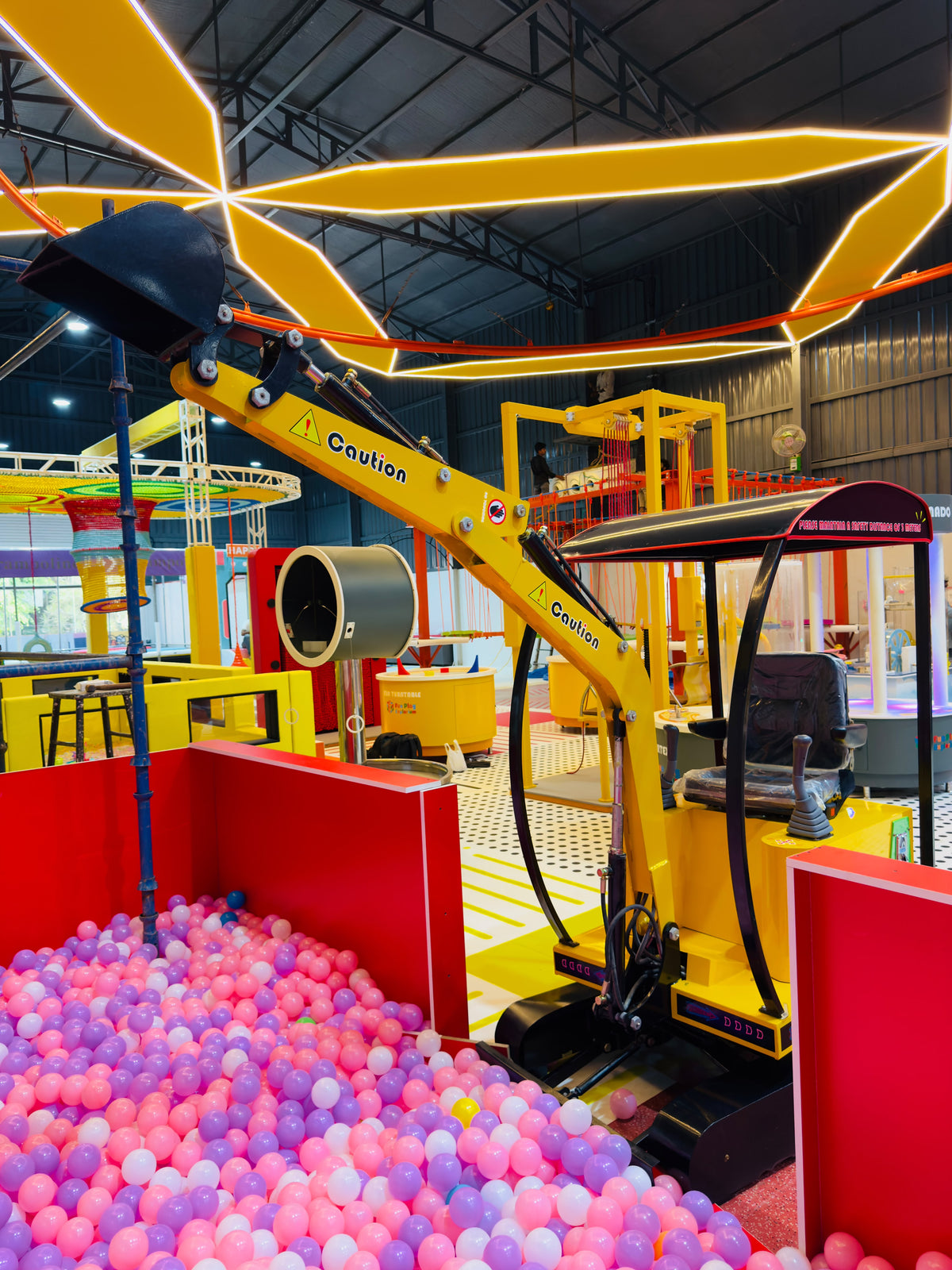 A yellow toy excavator sits in a ball pit filled with pink and purple balls.