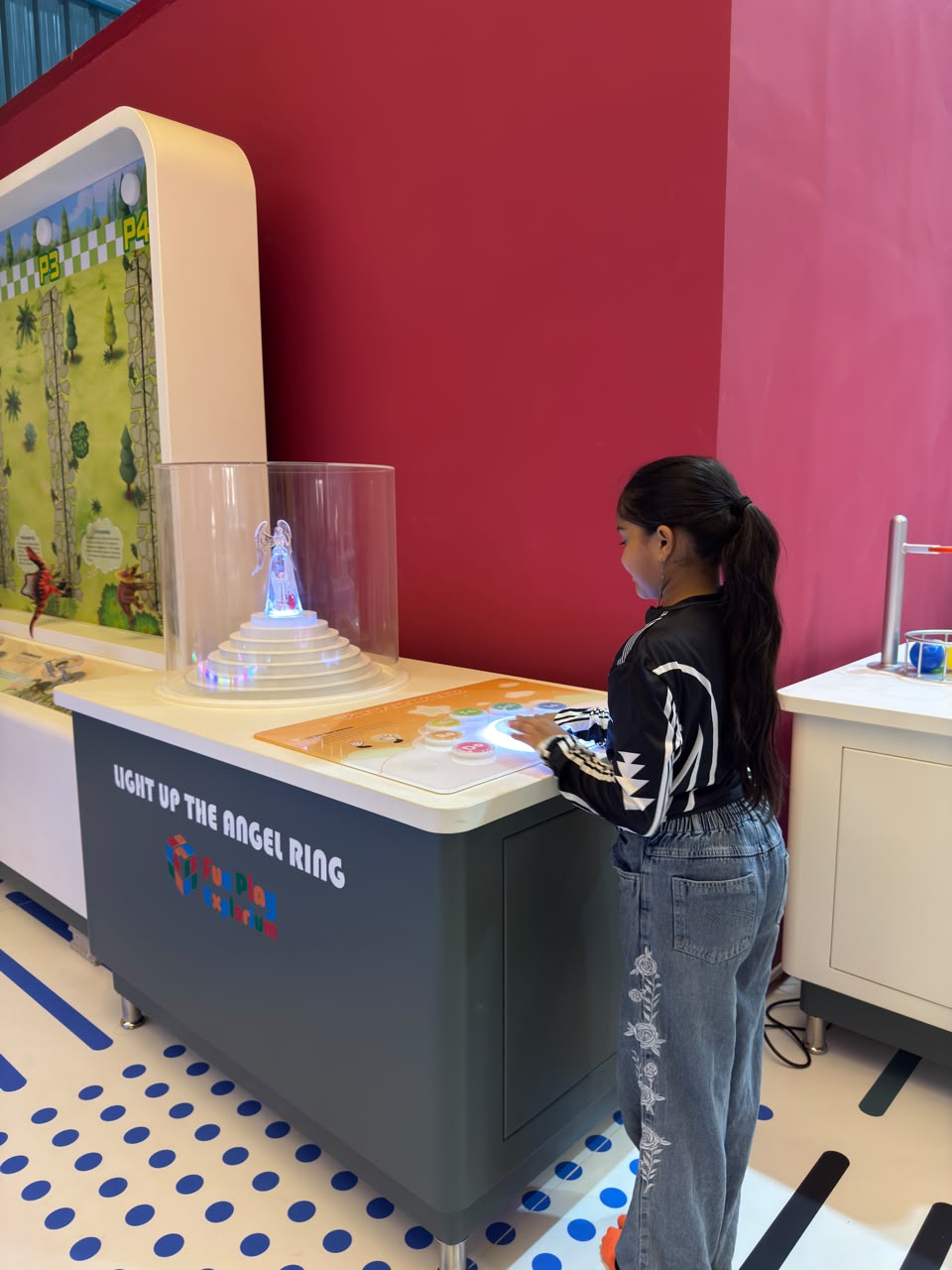 A young girl interacts with a colorful, illuminated game at an interactive exhibit. The game features a glowing angel figure and touch-sensitive buttons.