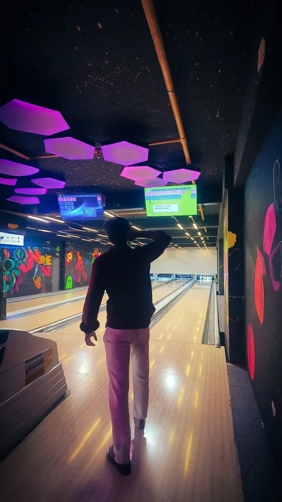 A person stands on a bowling lane, looking down the alley. The bowling alley has colorful murals and purple hexagonal lights on the ceiling.