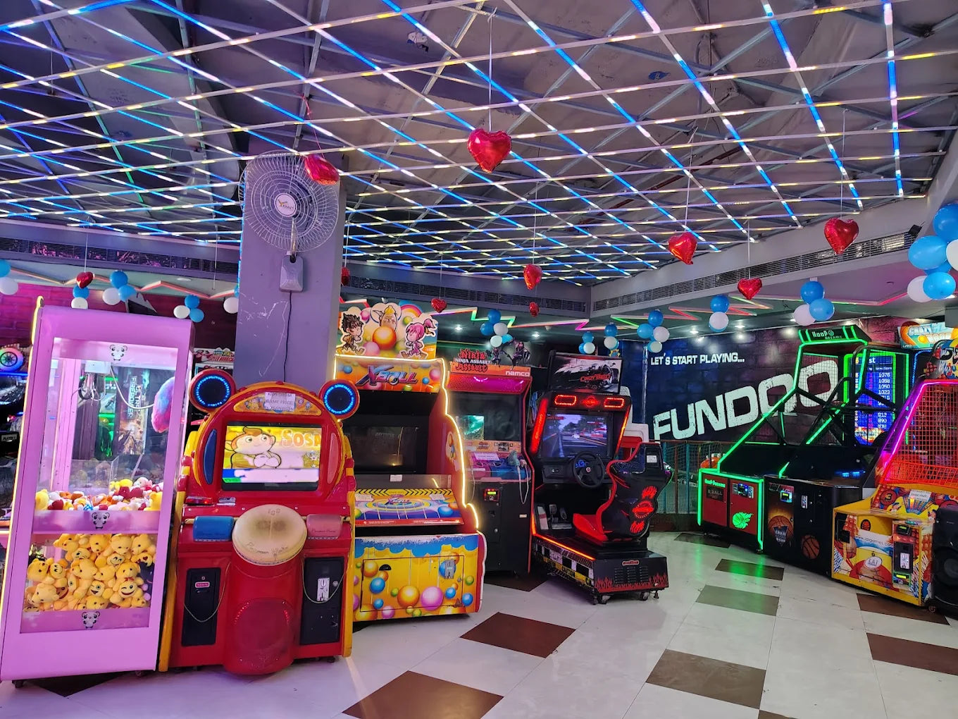 Indoor gaming arcade with bright machines and heart-shaped balloons under a glowing grid ceiling at Fundoo Gaming Zone Rohini