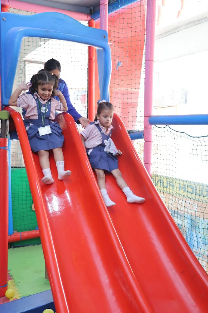 Two children sliding down a red slide at an indoor playground.