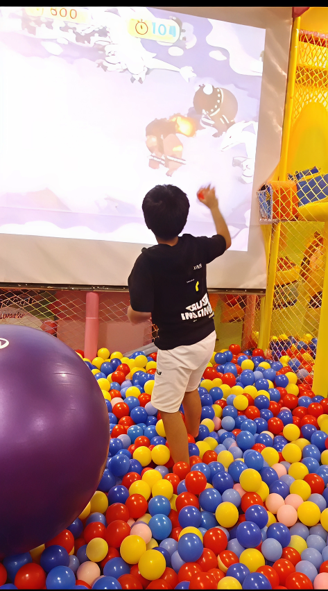 Child playing with colorful balls in a ball pit next to an interactive screen.
