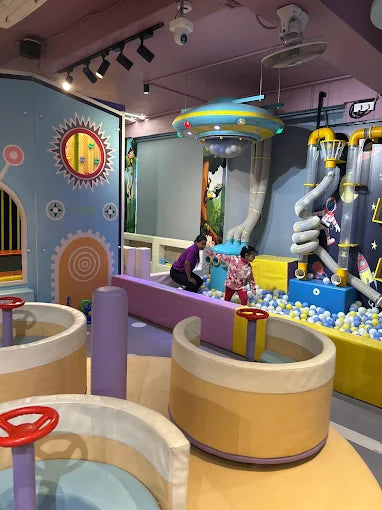 Children play in a colorful indoor playground with a ball pit and interactive elements. A large UFO-like structure hangs from the ceiling.