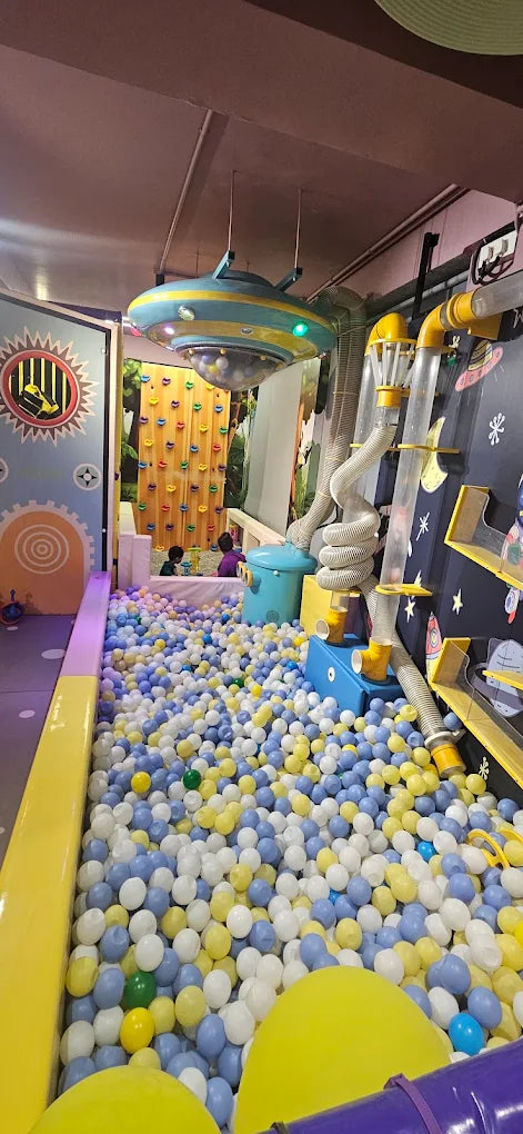 Children play in a ball pit with a climbing wall and UFO-themed decor.
