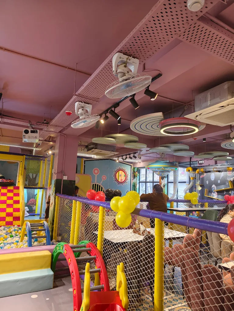 A colorful indoor playground with children playing in a ball pit and on slides, with adults seated at tables in the background.
