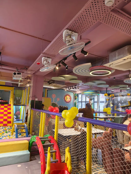 A colorful indoor playground with children playing in a ball pit and on slides, with adults seated at tables in the background.