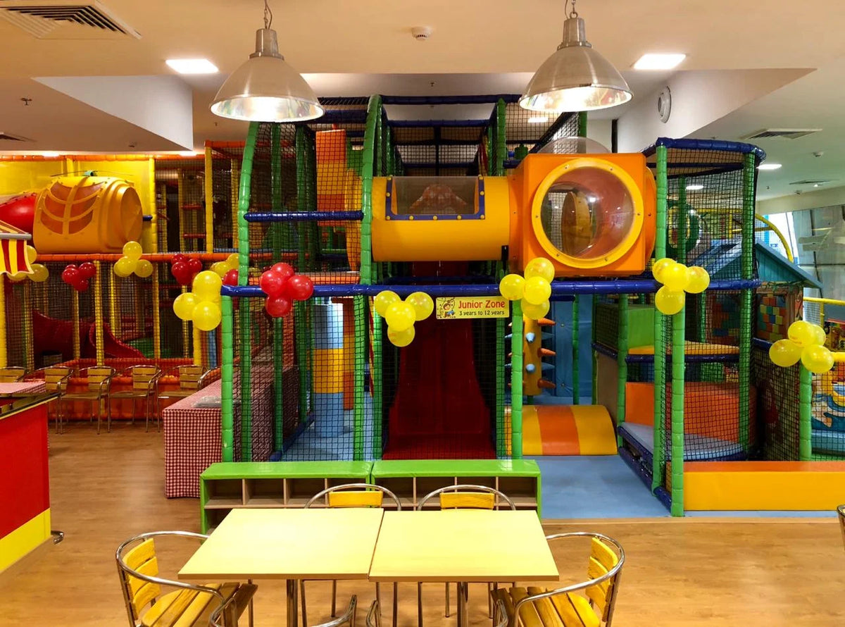 A colorful indoor playground with slides and tunnels, decorated with balloons, and tables and chairs in the foreground.
