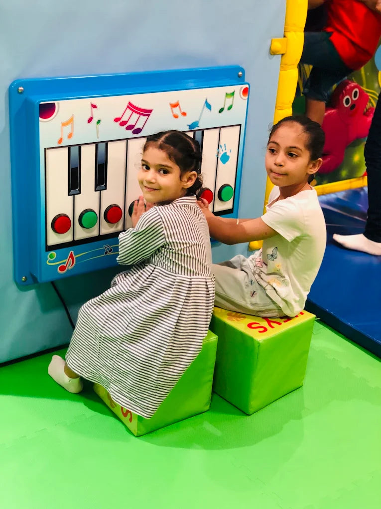 Two young girls play a large, colorful toy piano in a bright indoor play area.
