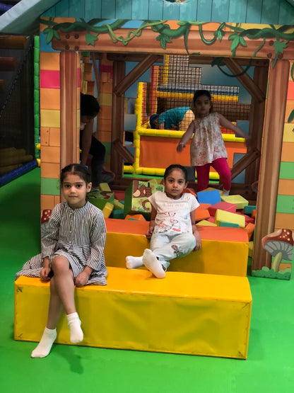 Two young girls sit on a yellow bench in a colorful indoor playground. Another girl stands behind them.