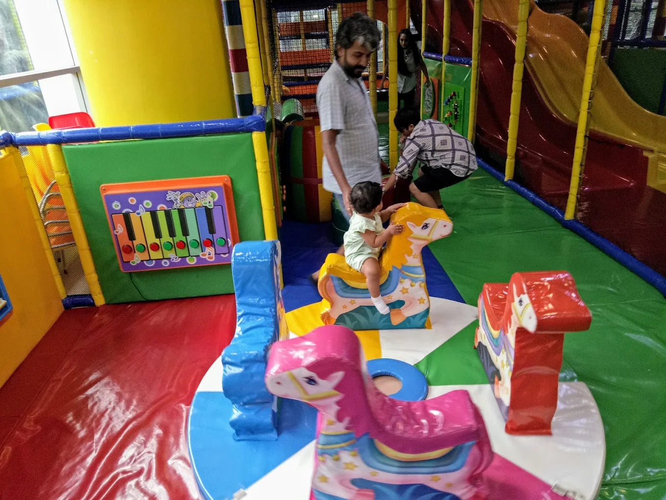A toddler rides a toy horse in a colorful indoor playground while adults watch.