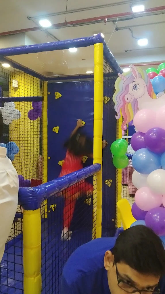 A young child climbs a colorful indoor playground wall, surrounded by balloons and a unicorn decoration.