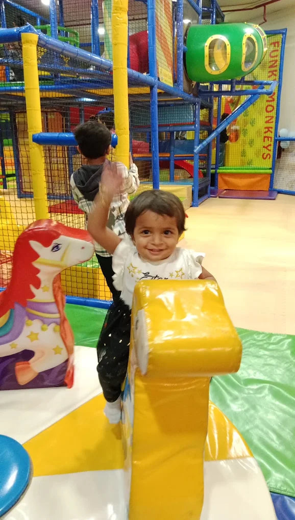 A toddler waves while sitting on a yellow ride at an indoor playground. Another child is visible in the background.
