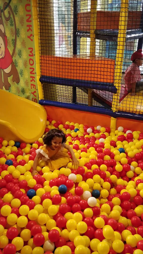 A young child plays in a ball pit next to a yellow slide in an indoor playground.